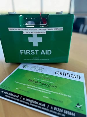 Green first aid kit with a white cross and the words 'First Aid' on it. Next to it is a certificate of completion for a 3-day First Aid at Work course issued by Aberdeen First Aid School to Holly Stewart.
